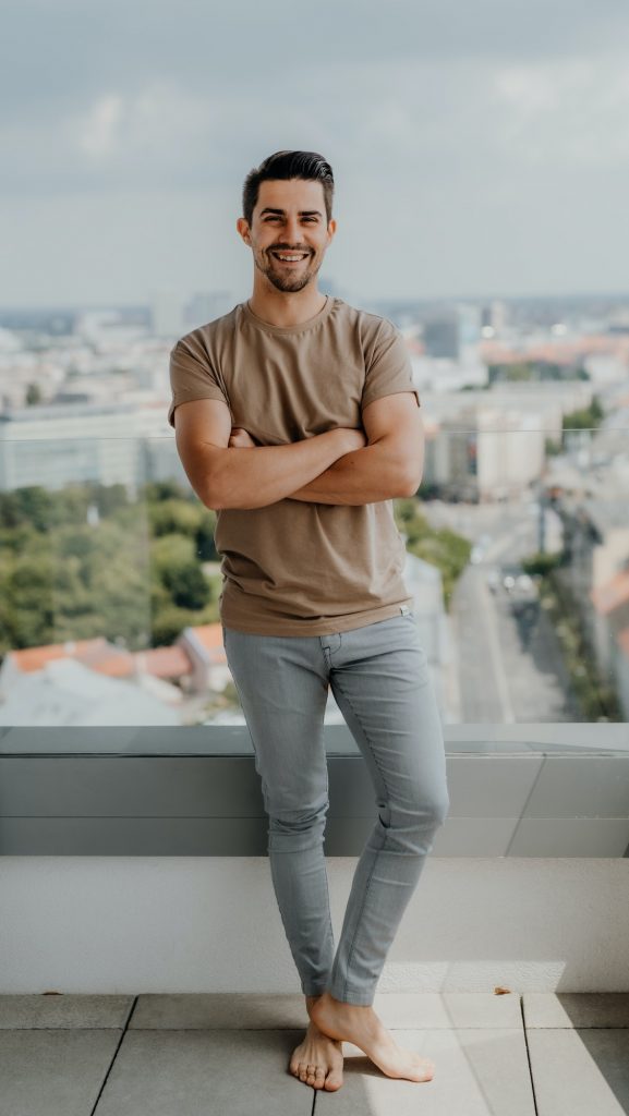 Portrait of happy young caucasian man with arms crossed standing on balcony, looking at camera